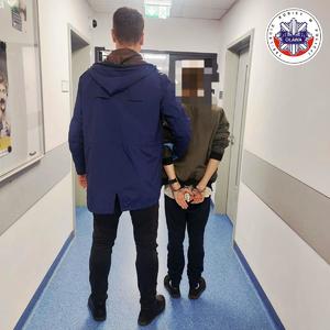 Policjant prowadzący po swojej prawej stronie zatrzymanego mężczyznę, który ma kajdanki założone na ręce trzymane z tyłu